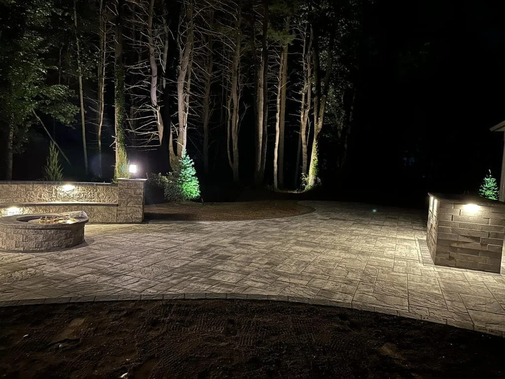 Stone patio with built-in fire pit and landscape lighting illuminating trees at night.