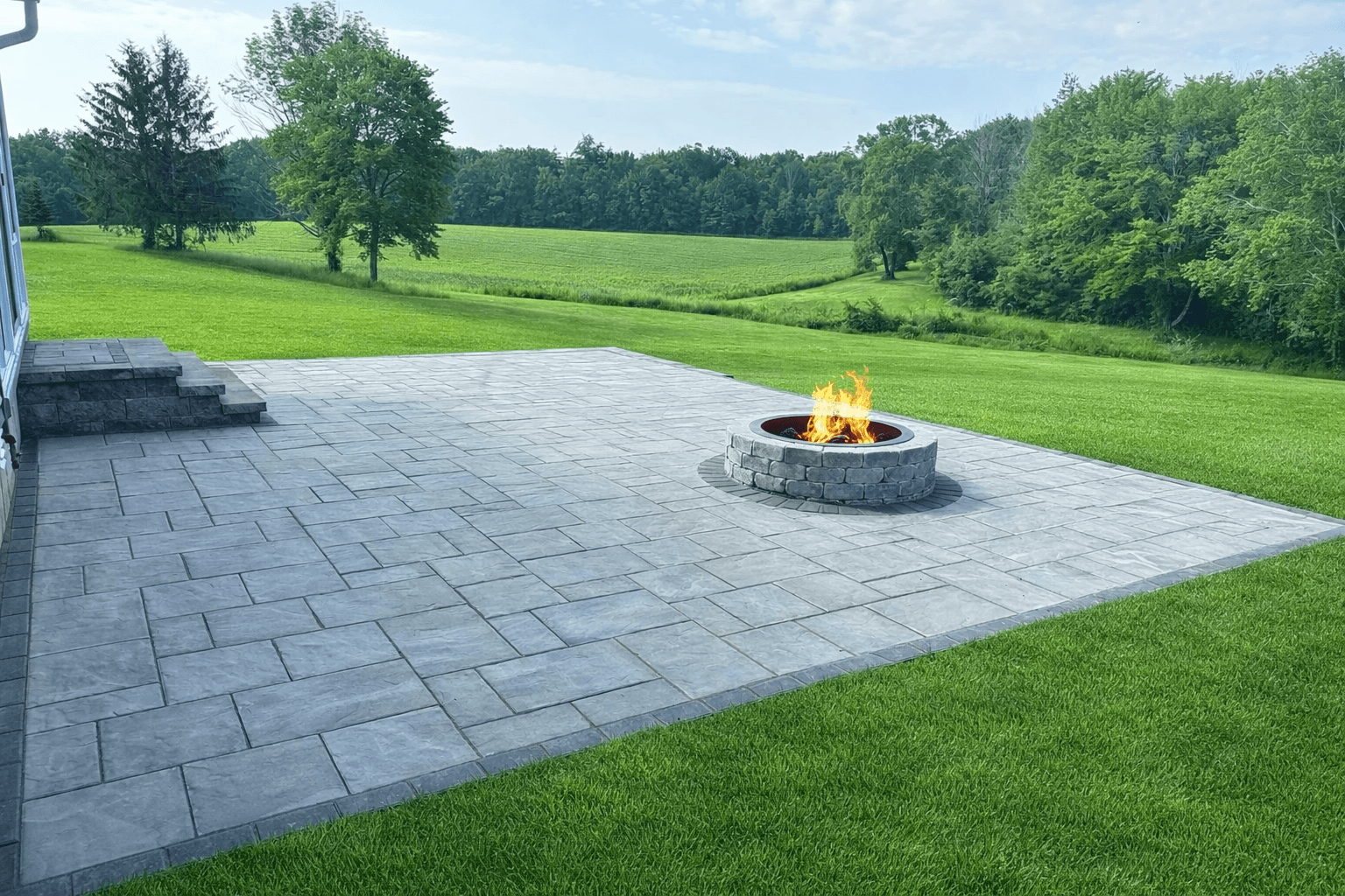 Large backyard paver patio with circular fire pit overlooking open green landscape