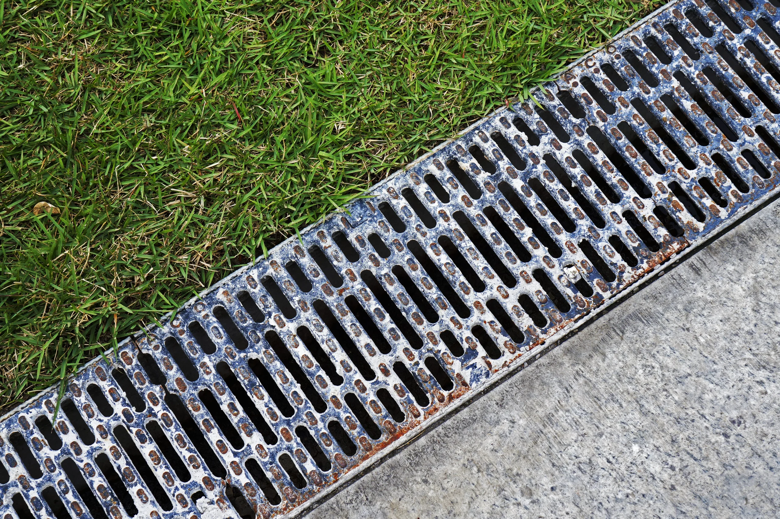 Metal drainage grate installed beside grass and concrete pavement to manage rainwater runoff.