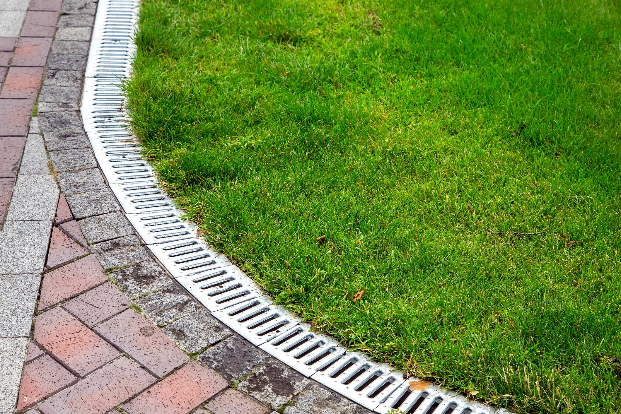 Close-up of a linear drainage grate separating green turf from a curved brick pathway for water runoff control.