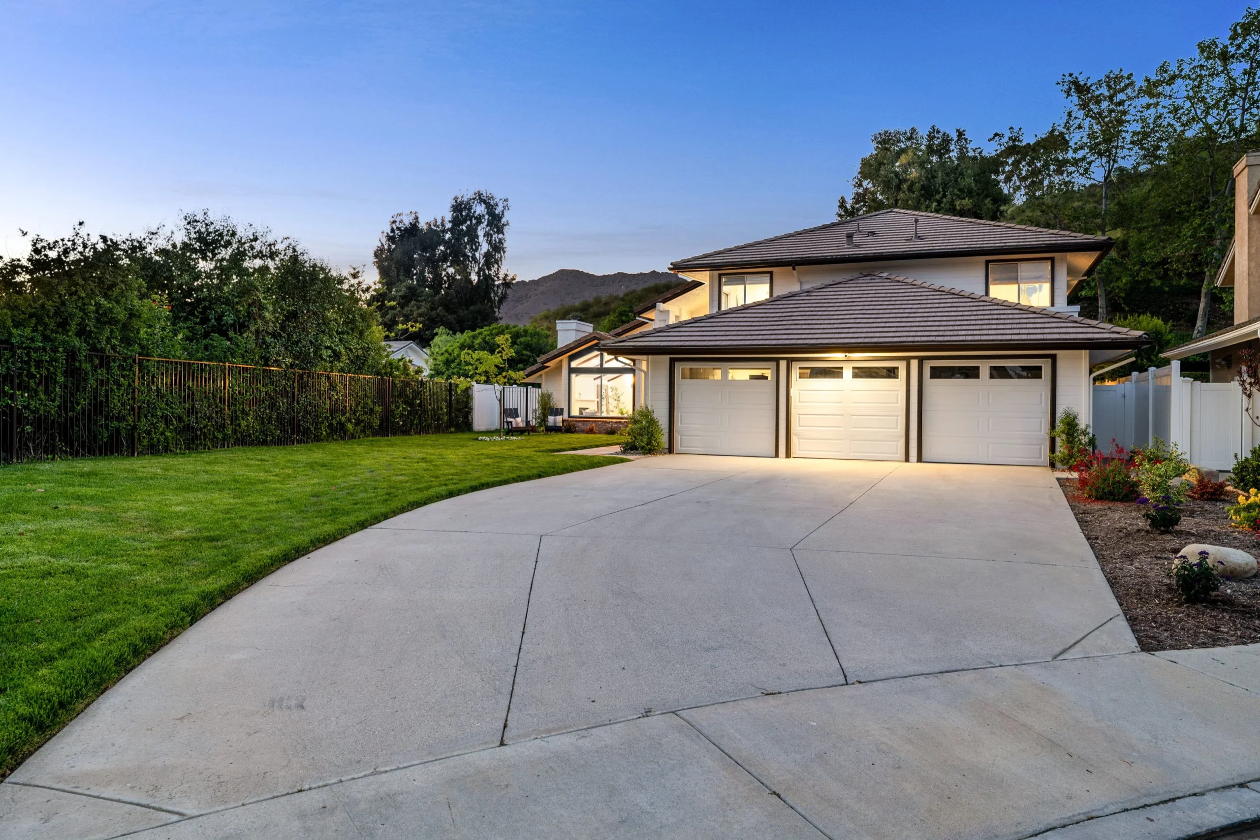 Modern suburban home featuring a broad concrete driveway, green lawn, and illuminated garage doors at dusk.