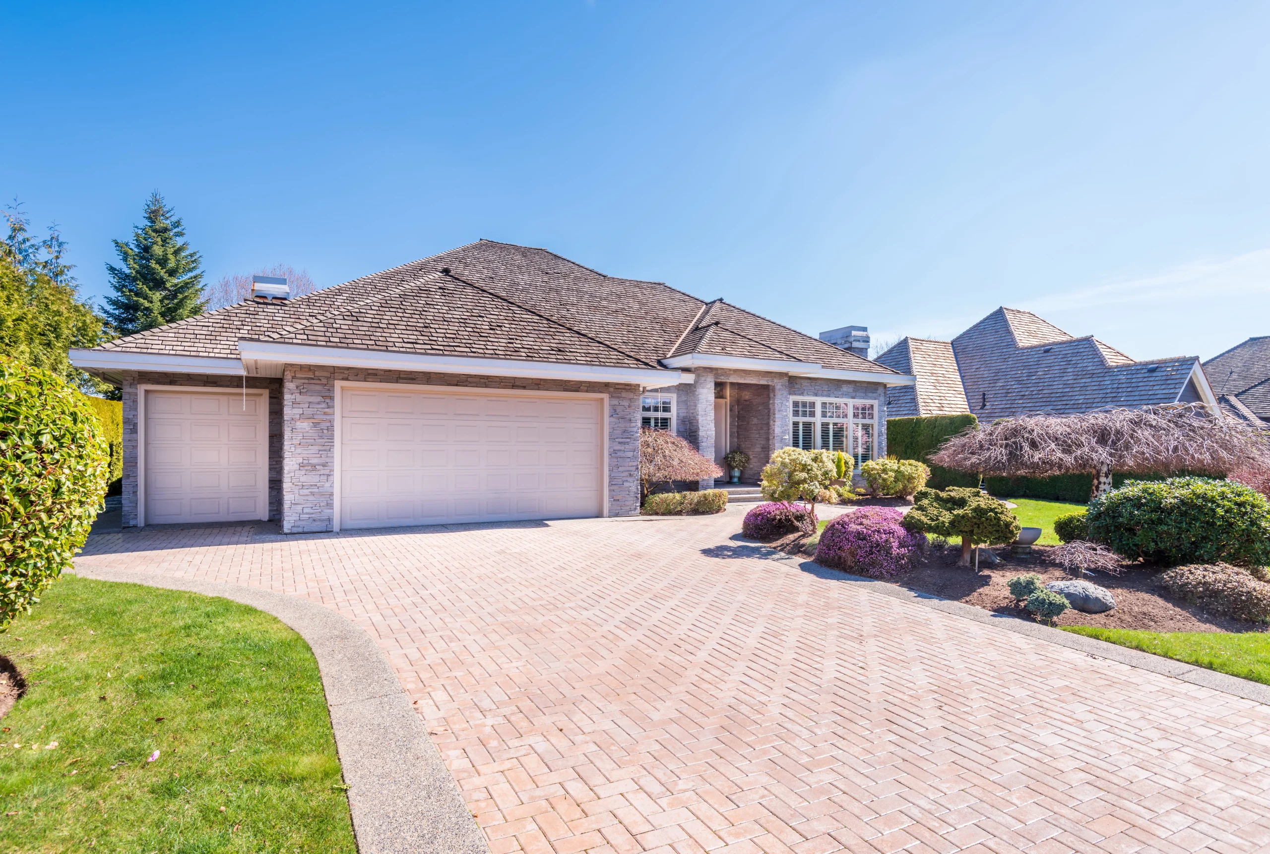 Modern suburban home featuring a wide brick driveway, manicured lawn, and decorative shrubs.