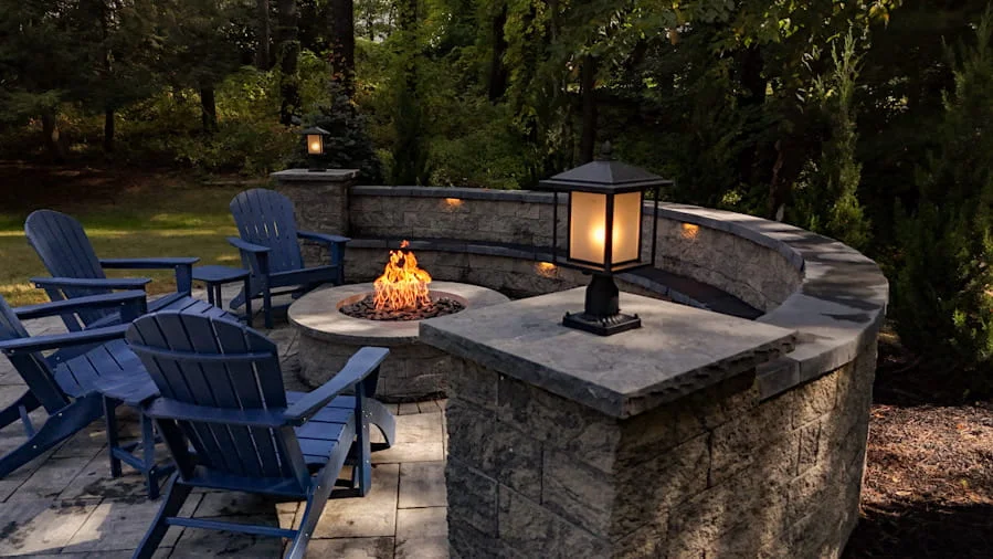 Backyard patio featuring round fire pit, blue chairs, stone seating wall, and landscape lighting.