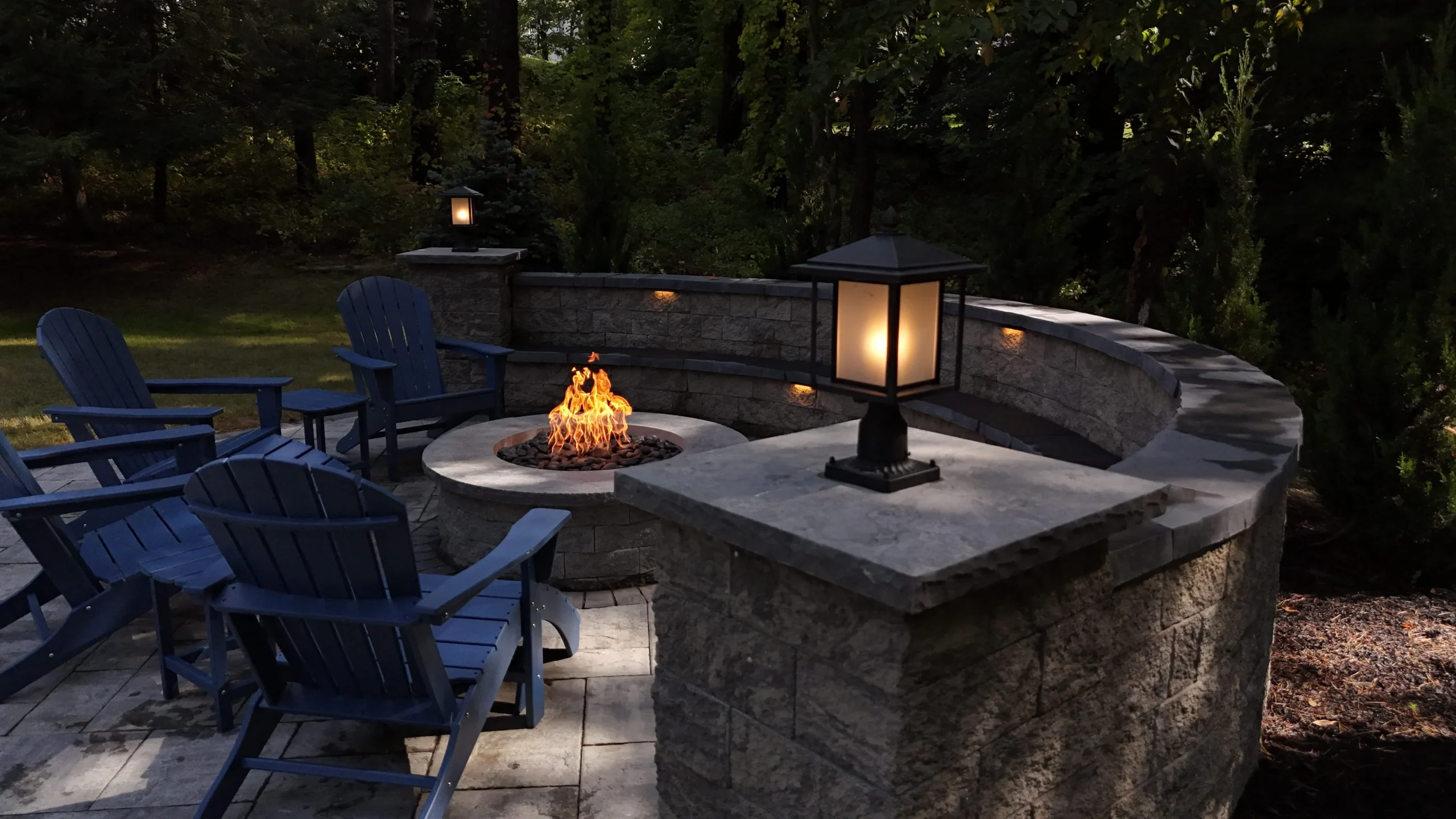 Stone patio with built-in fire pit, Adirondack chairs, and soft landscape lighting at night.