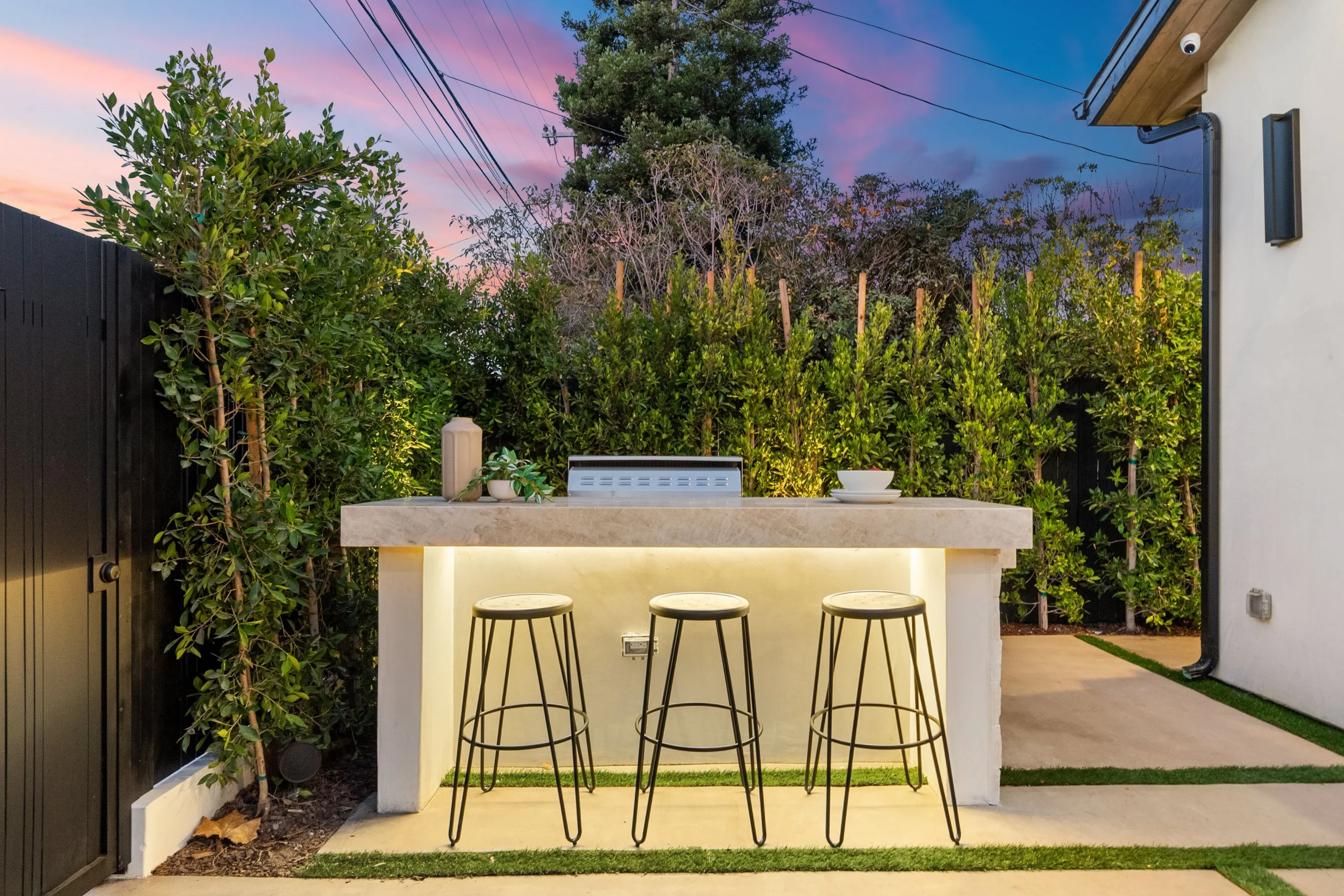 Modern outdoor bar counter with stools and built-in lighting in a landscaped backyard.