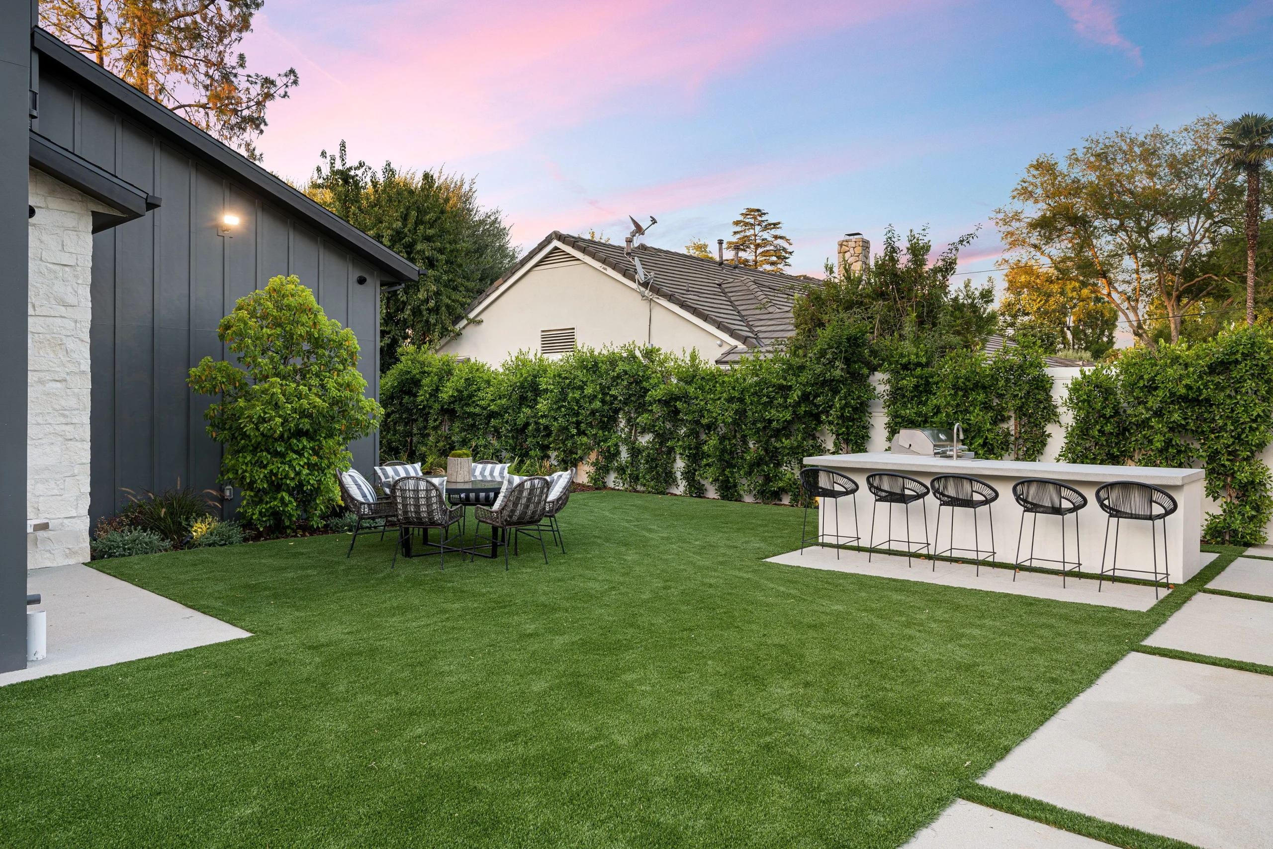 Backyard patio with outdoor bar seating, dining table, and landscaped lawn at sunset in New York.