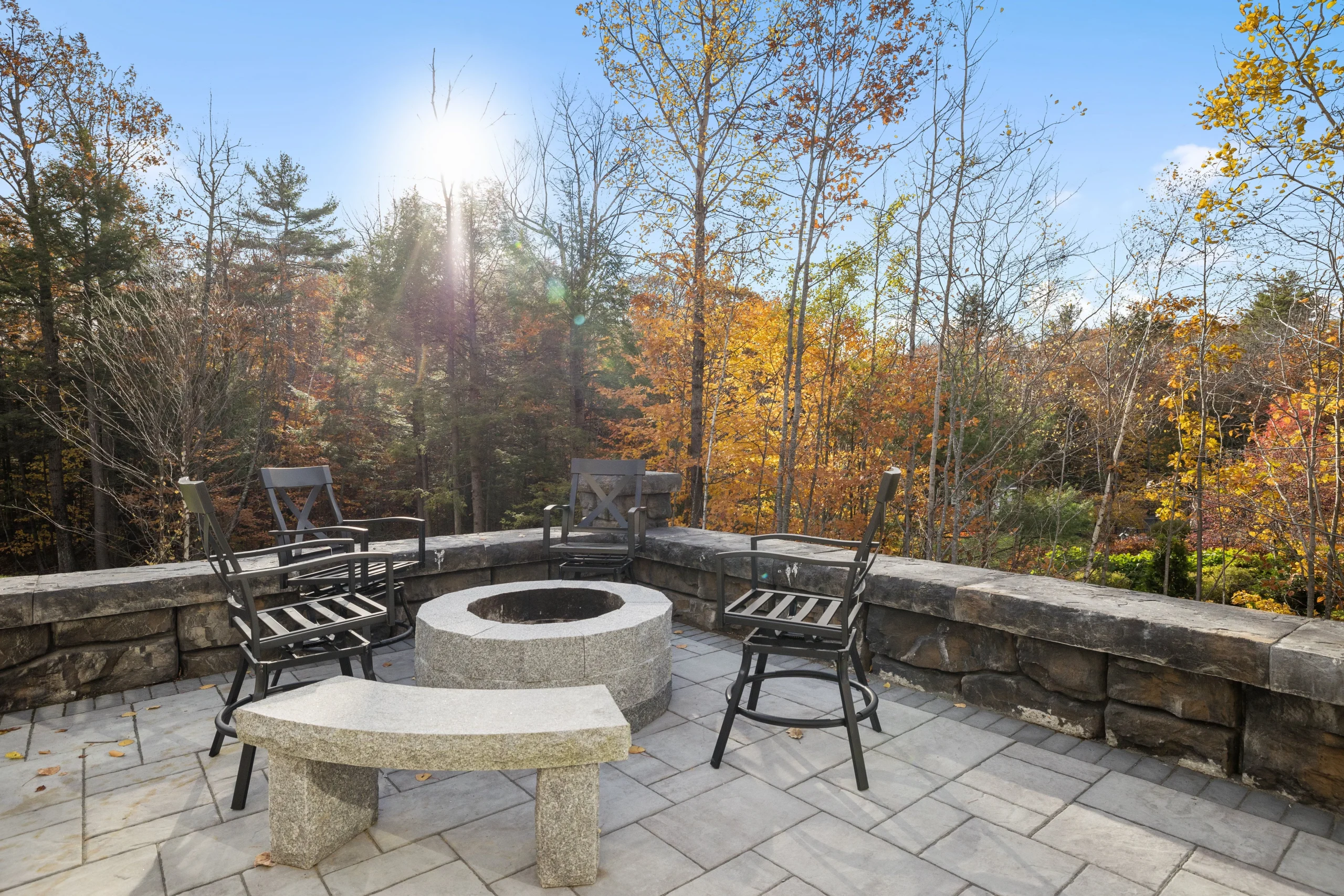 Patio fire pit surrounded by chairs and stone wall with scenic fall foliage view.