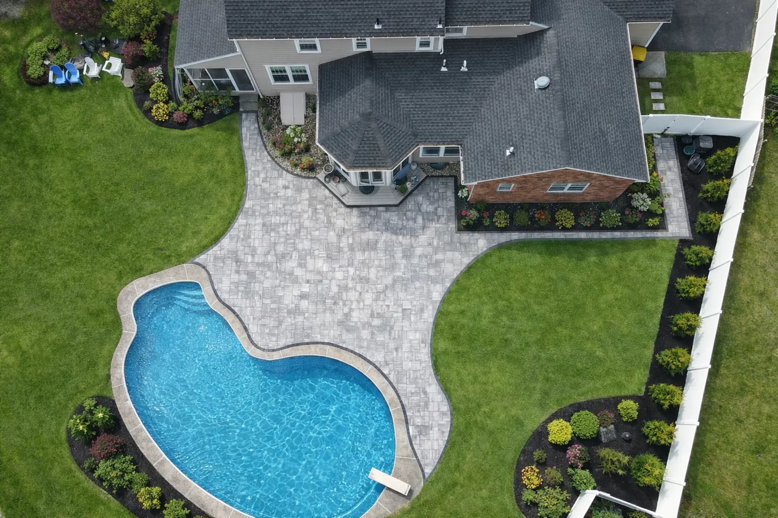 Drone view of a residential backyard featuring a curved swimming pool, stone paver patio, and landscaped lawn.